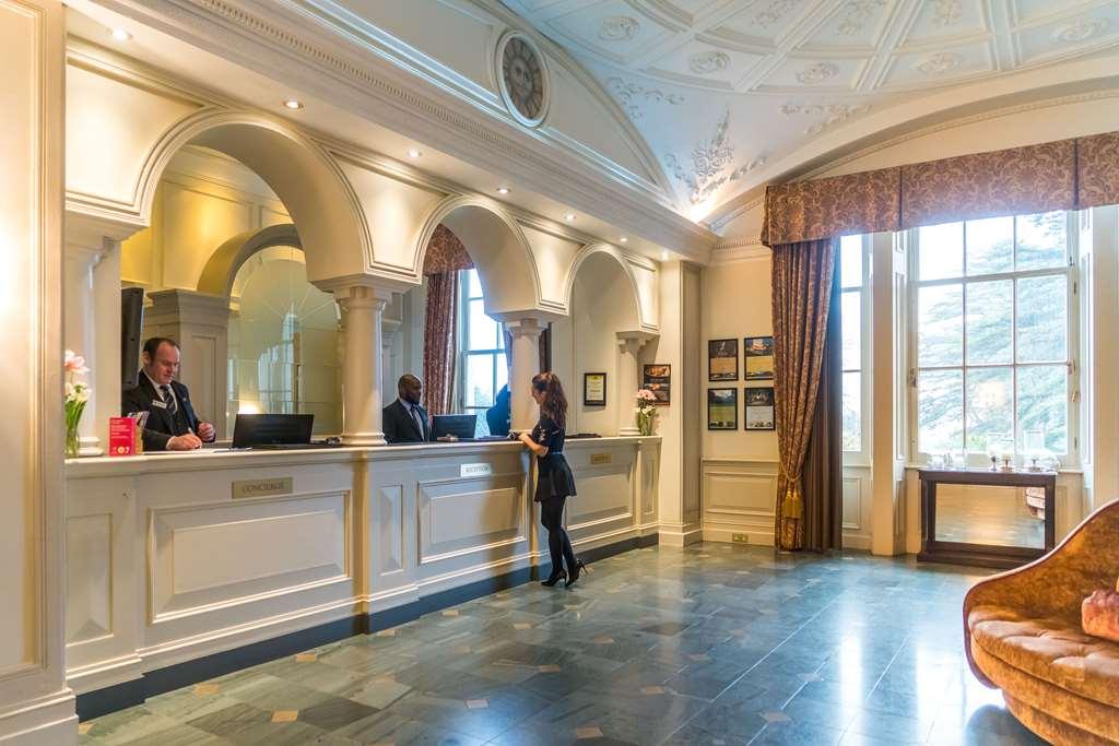 맥도날드 배스 스파 호텔 바스 내부 사진 The photo shows the interior of a hotel reception area. There is a stylish front desk with several staff members attending to guests. A woman is interacting with one of the receptionists. The design features elegant architectural details, including a