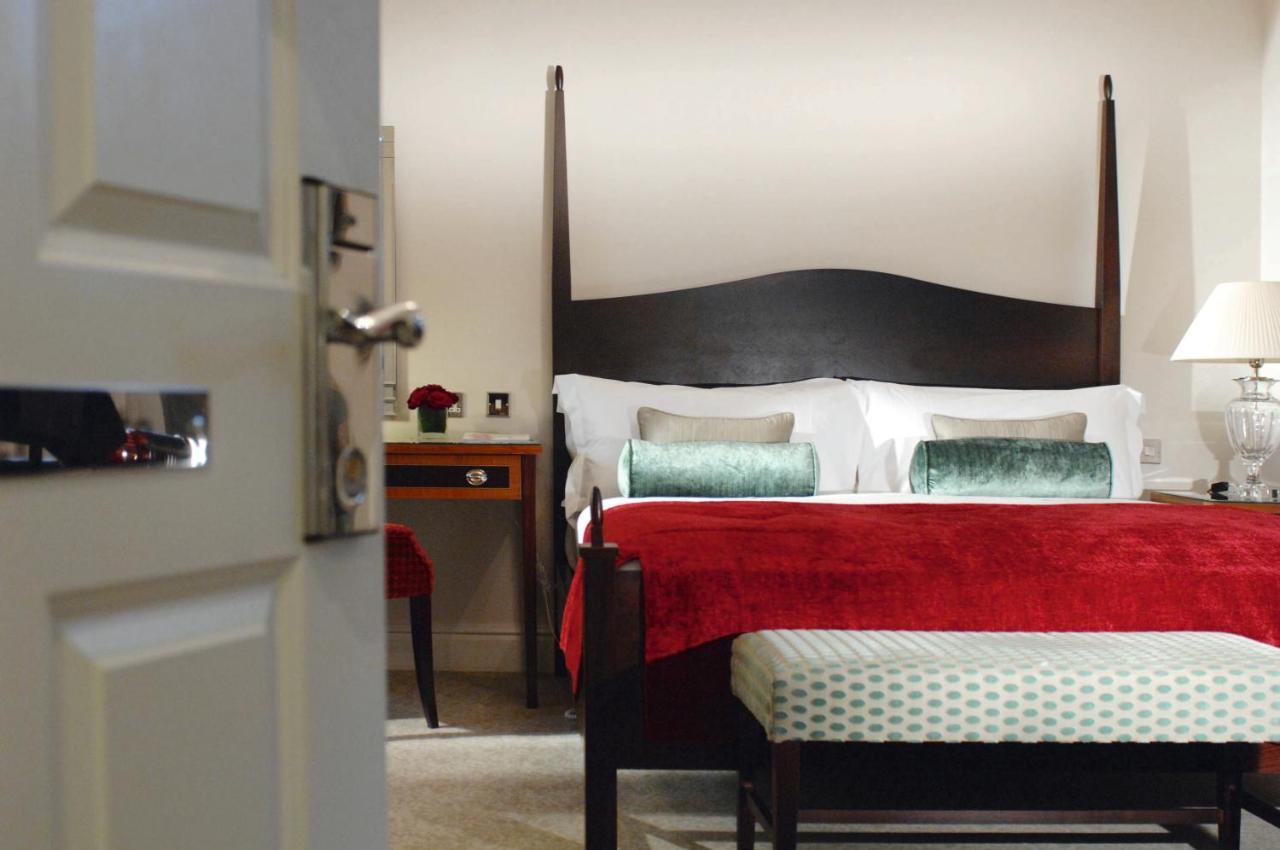 맥도날드 배스 스파 호텔 바스 객실 사진 The photo shows a well-decorated hotel room. In the foreground, there is a partially opened door leading into the room. The focal point of the room is a large bed with a dark wooden headboard and stylish red and white bedding, accented with green pil