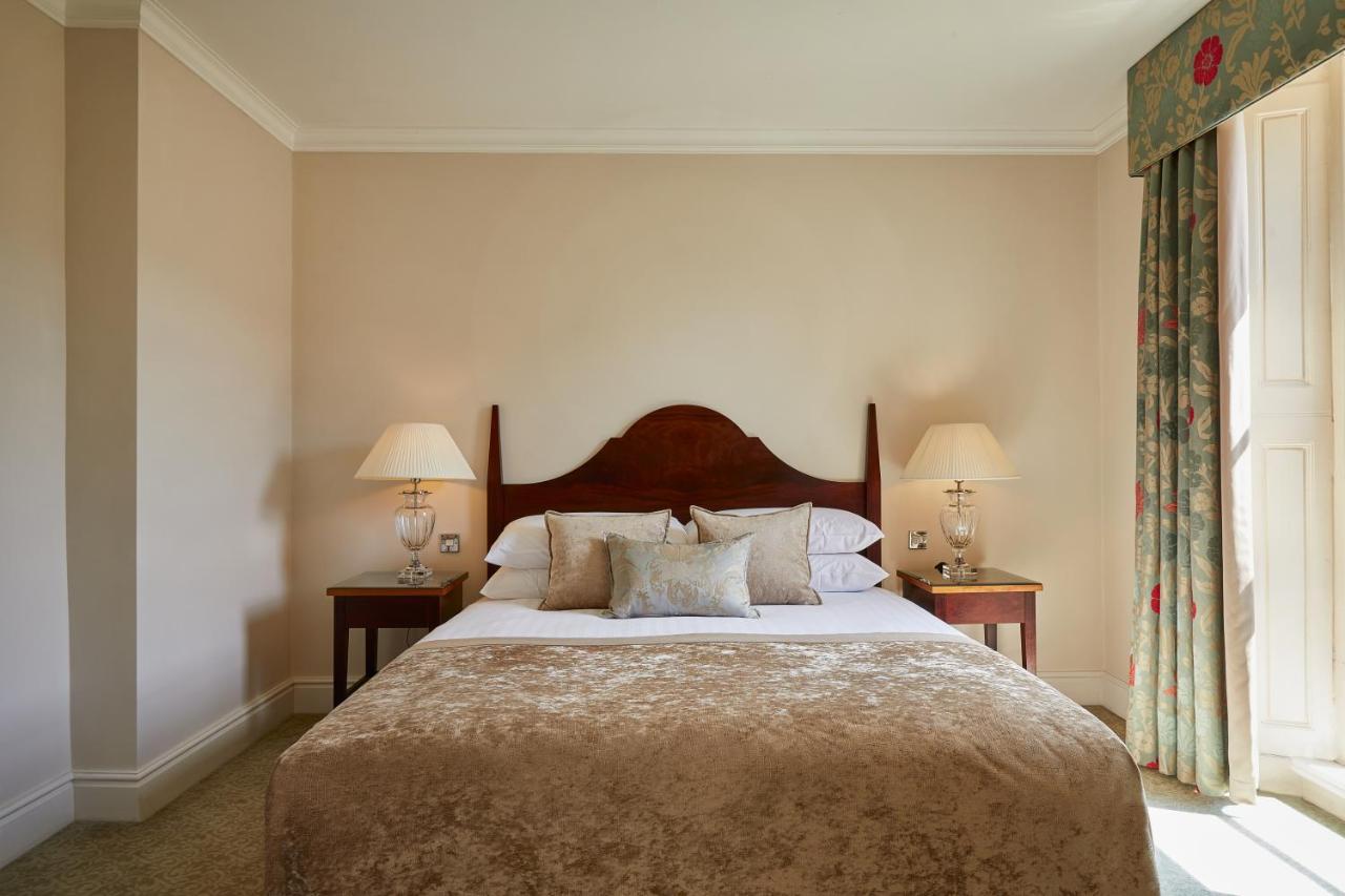 맥도날드 배스 스파 호텔 바스 외부 사진 The photo shows a cozy hotel bedroom. It features a large bed with a plush, light-colored bedspread and several pillows. On either side of the bed, there are wooden nightstands with lamps providing soft lighting. The walls are painted a neutral color