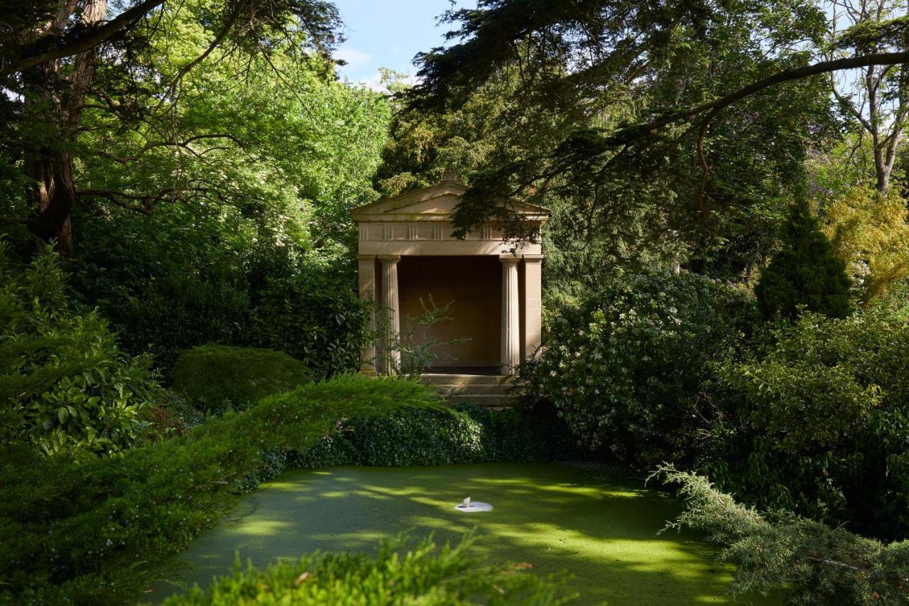 맥도날드 배스 스파 호텔 바스 외부 사진 The photo shows a serene garden scene featuring lush greenery and a tranquil pond. In the background, there is a classical-looking structure with columns, resembling a small temple or pavilion. The pond appears to have vegetation covering its surface