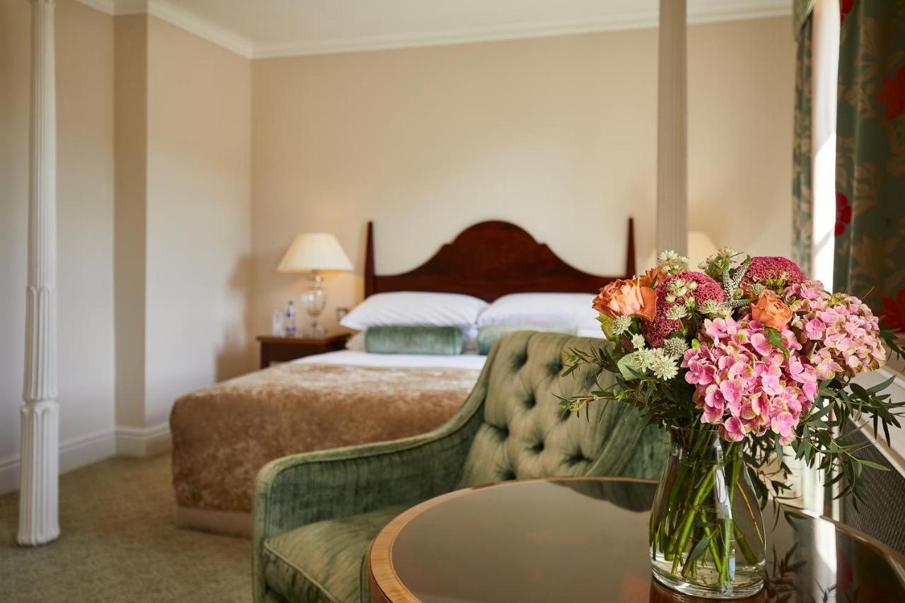맥도날드 배스 스파 호텔 바스 외부 사진 The photo shows a cozy bedroom featuring a large bed with a wooden headboard, topped with a fluffy blanket. To one side, there is a small bedside lamp providing soft lighting. In the foreground, there is an elegant green chair with a tufted design an