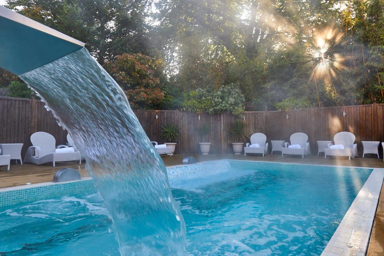 맥도날드 배스 스파 호텔 바스 외부 사진 The photo shows a tranquil swimming pool surrounded by greenery. Water is cascading from a feature into the pool, creating a refreshing effect. In the background, there are white lounge chairs positioned neatly along the poolside, inviting relaxation