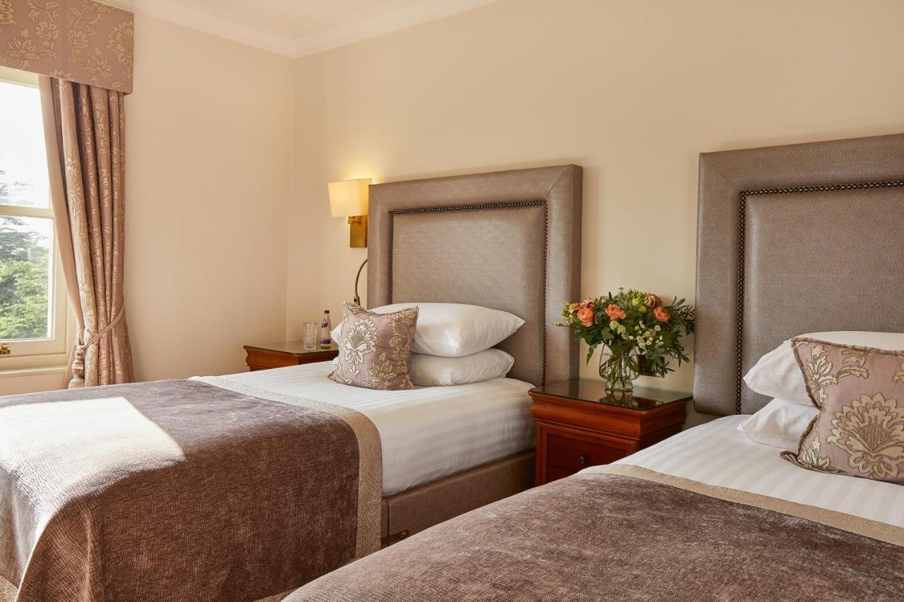 맥도날드 배스 스파 호텔 바스 외부 사진 The photo shows a hotel room featuring two beds with elegant headboards. Each bed is neatly made with white linens and a soft, decorative throw blanket. There are matching pillows on the beds, and a small, stylish bouquet of flowers is placed on a wo