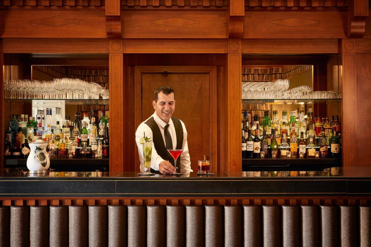 맥도날드 배스 스파 호텔 바스 외부 사진 The photo depicts a bartender standing behind a polished bar. He is wearing a formal outfit with a vest and a bow tie, and he appears to be smiling as he presents a cocktail. Behind him is a well-stocked bar shelf filled with various bottles of liquo