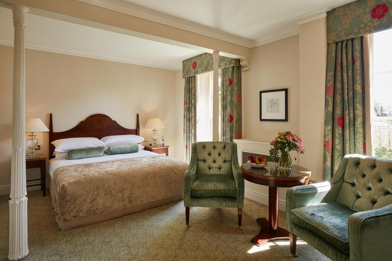 맥도날드 배스 스파 호텔 바스 외부 사진 The photo shows a cozy hotel room featuring a large bed with a beige bedspread. There are two green armchairs positioned next to a small round table. The decor includes floral curtains and a neutral color palette on the walls. Soft lighting is provid