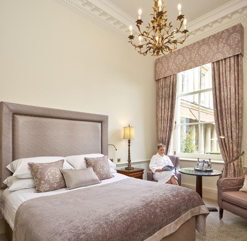 맥도날드 배스 스파 호텔 바스 외부 사진 The photo shows a well-appointed hotel room that features a large, elegant bed with soft bedding and decorative pillows. The walls are painted a light color, and there is an ornate chandelier hanging from the ceiling. A beautiful window is adorned wi
