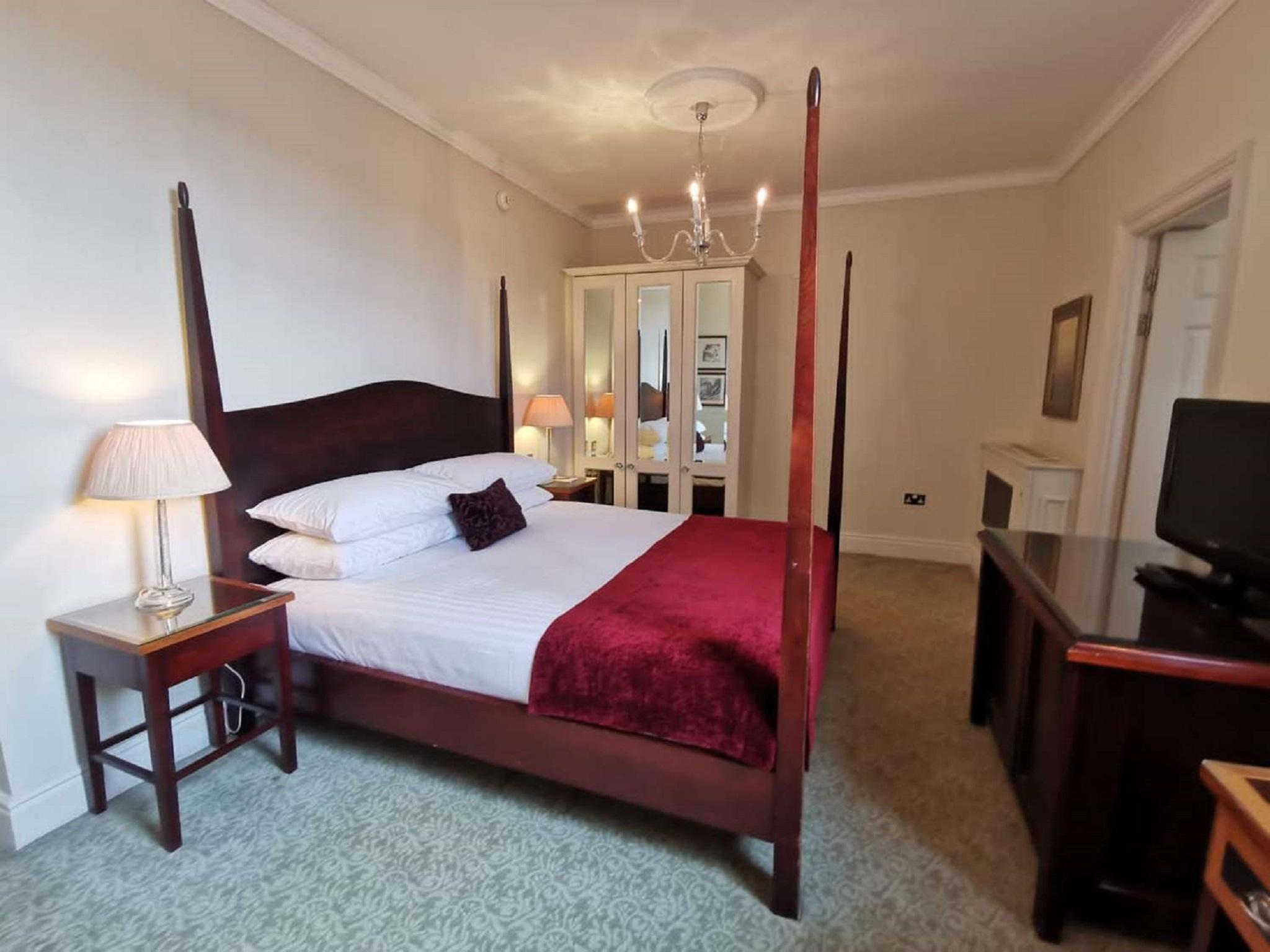 맥도날드 배스 스파 호텔 바스 외부 사진 The photo shows a well-decorated bedroom featuring a four-poster bed with white pillows and a burgundy throw. There are two bedside lamps on wooden nightstands on either side of the bed. A large wardrobe or mirror cabinet is visible in the background