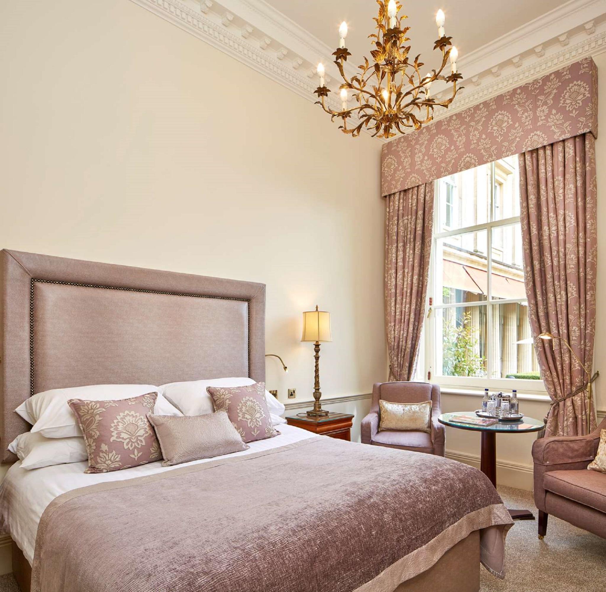 맥도날드 배스 스파 호텔 바스 외부 사진 The photo shows a beautifully decorated hotel room. It features a large bed with a plush headboard, adorned with several decorative pillows. The walls are painted in a soft color, and there is a chandelier hanging from the ceiling. Natural light come