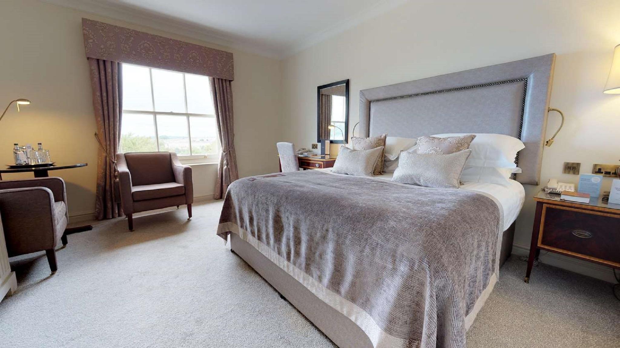 맥도날드 배스 스파 호텔 바스 외부 사진 The photo shows a well-furnished bedroom. It features a large bed with a light gray headboard and several decorative pillows on top. The bed is covered with a soft gray blanket. To one side of the room, there is a comfortable armchair and a small tab