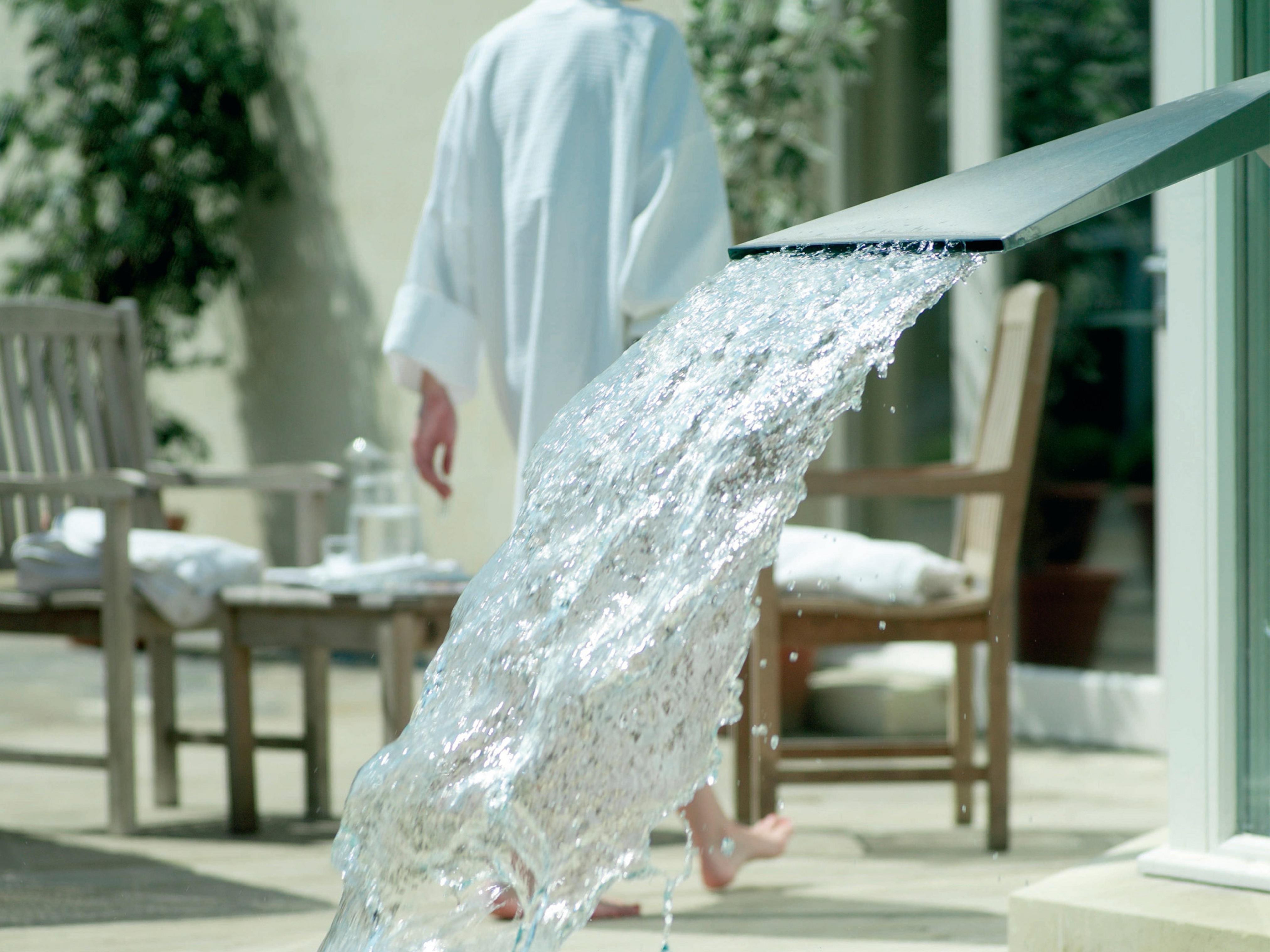 맥도날드 배스 스파 호텔 바스 외부 사진 The photo shows a flowing water feature, with water elegantly cascading from a sleek spout. In the background, there is a person wearing a white bathrobe, walking away. Surrounding the area, there are wooden chairs and what appears to be a serene out