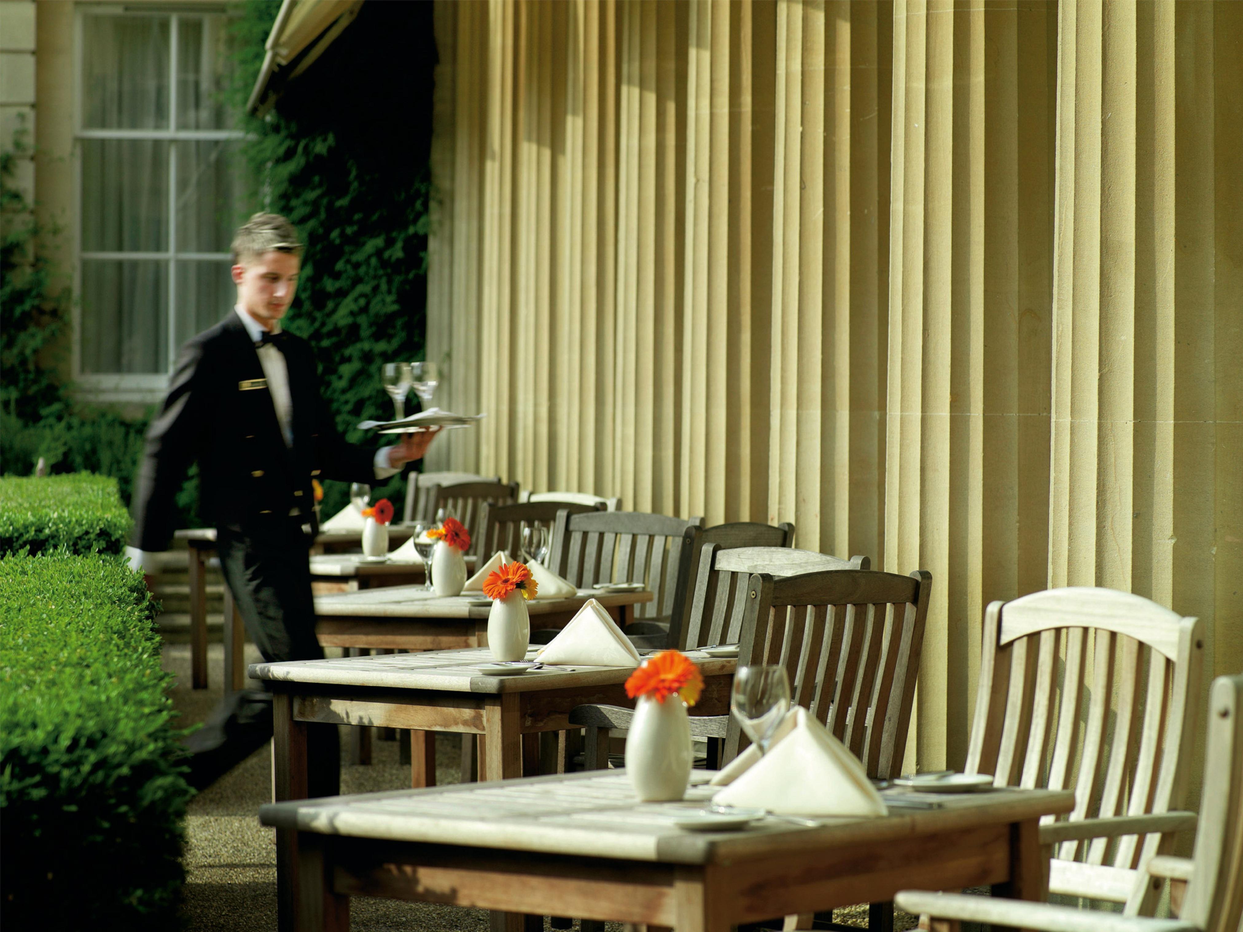 맥도날드 배스 스파 호텔 바스 외부 사진 The photo shows an outdoor dining area, featuring several wooden tables and chairs arranged neatly. Each table is set with a white tablecloth and small vases containing flowers, adding a vibrant touch. A waiter in a formal black suit is seen moving t