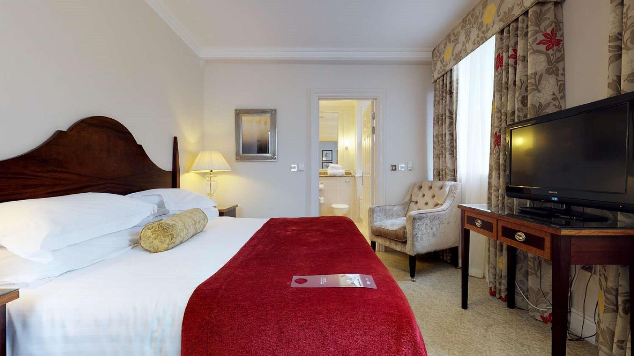 맥도날드 배스 스파 호텔 바스 외부 사진 The photo shows a hotel room with a queen-sized bed covered in white linens and a deep red throw blanket. There is a decorative pillow on the bed. Next to the bed, there's a small nightstand with a lamp. In the background, a seating area features a b