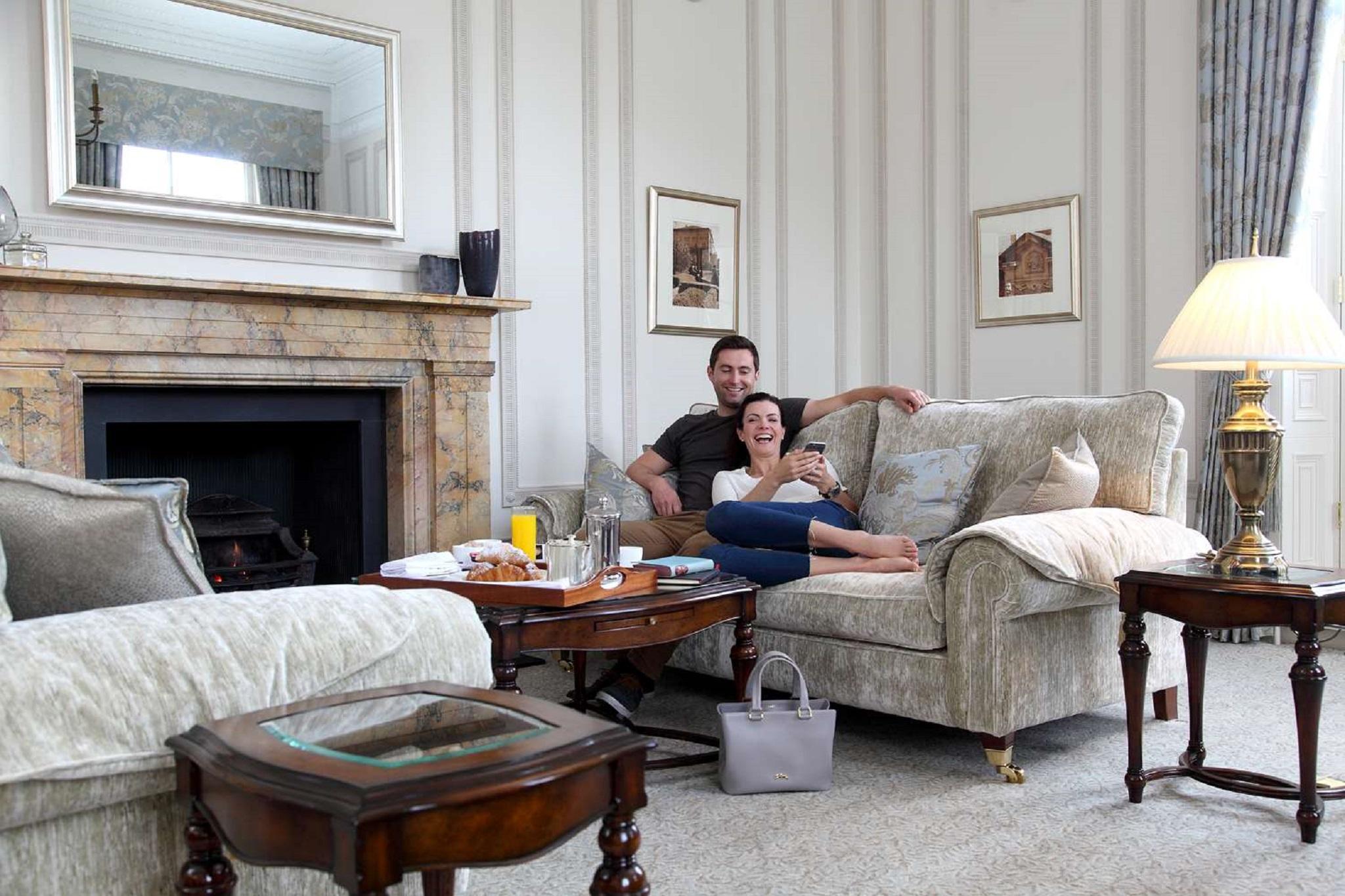 맥도날드 배스 스파 호텔 바스 외부 사진 The photo shows a cozy living room setting where a couple is sitting together on a light-colored sofa. They appear relaxed, with the man sitting slightly behind the woman, who is smiling and looking at her phone. The room features elegant decor, incl