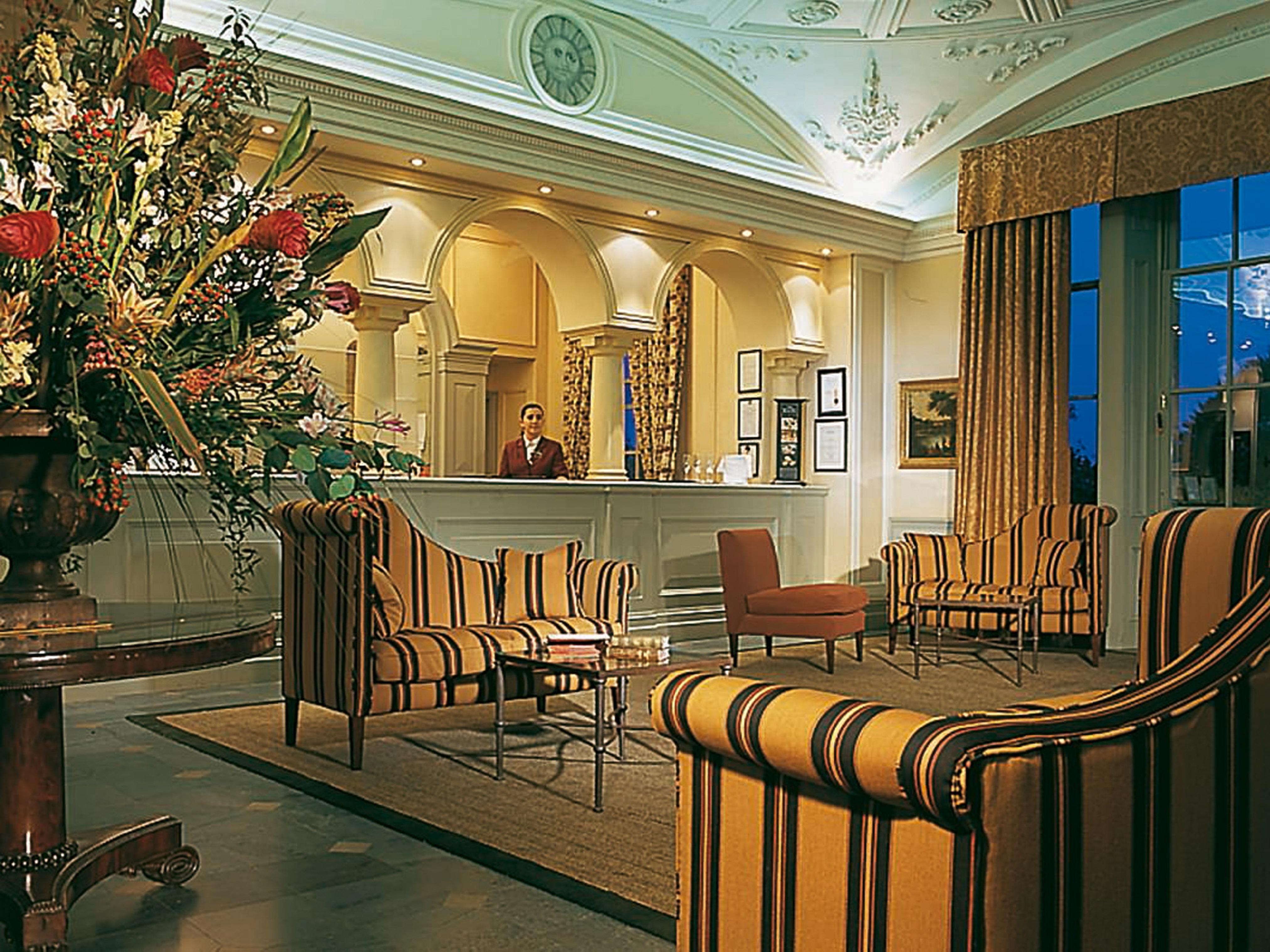 맥도날드 배스 스파 호텔 바스 외부 사진 The photo shows an elegant hotel lobby. The area features a reception desk with a staff member behind it. Surrounding the reception are comfortable seating arrangements, including striped armchairs and a brown chair. A large vase with an elaborate fl
