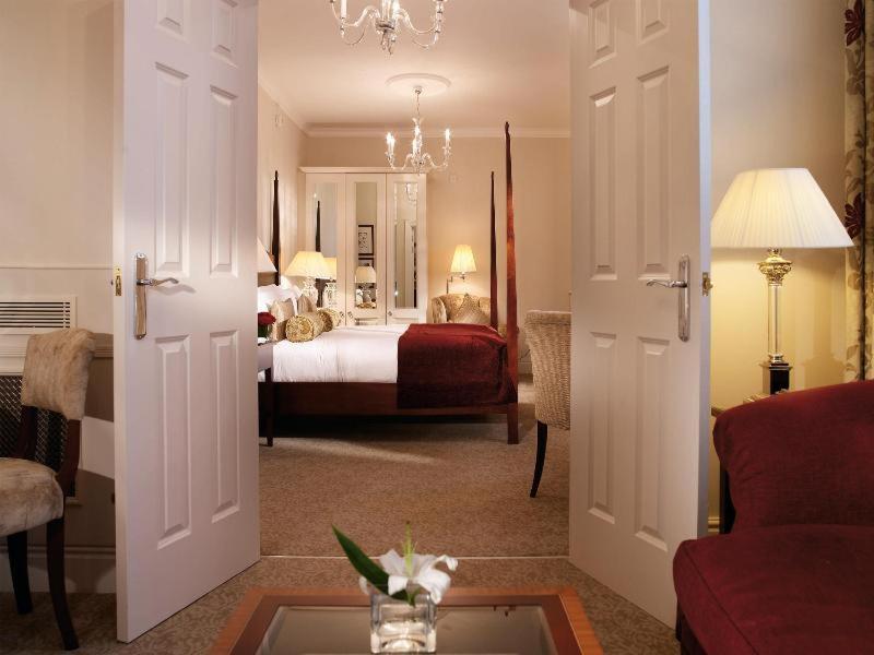 맥도날드 배스 스파 호텔 바스 객실 사진 The photo shows a hotel room that can be viewed through open double doors. Inside, there is a neatly made bed with decorative pillows, a plush area rug, and soft lighting from lamps. The room features elegant furnishings, including a chair, a coffee 