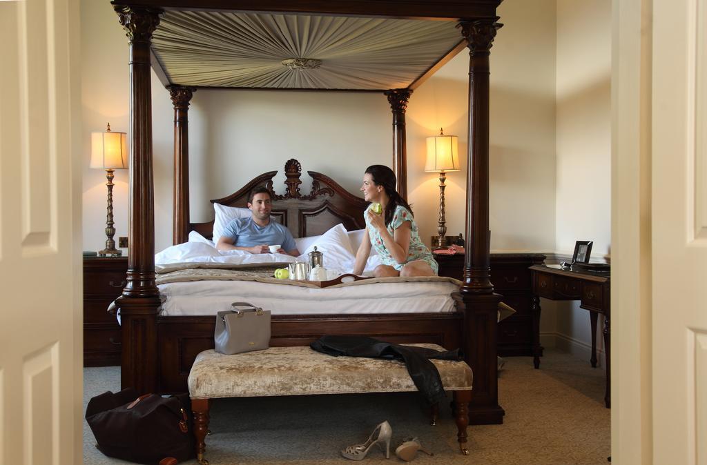 맥도날드 배스 스파 호텔 바스 외부 사진 The image depicts a spacious, well-lit bedroom featuring a large four-poster bed with an ornate headboard. A man and a woman are sitting on the bed, appearing to engage in conversation. The woman is dressed in a patterned outfit, while the man is wea