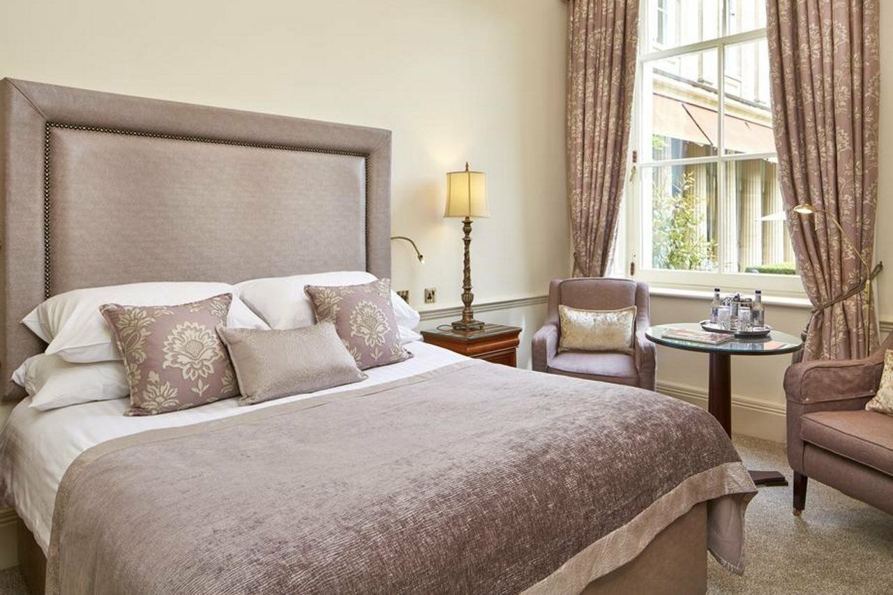 맥도날드 배스 스파 호텔 바스 외부 사진 The photo shows a stylish and elegantly decorated hotel room. It features a large bed with soft linens and several decorative pillows. The bedspread is a muted shade of beige or taupe. Beside the bed, there's a small table with two armchairs. One of 